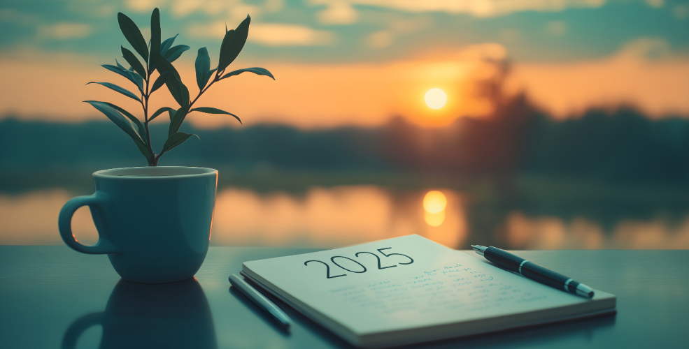 A serene scene of a notebook labeled "2025," accompanied by a pen and a cup with a growing plant, set against the backdrop of a golden sunrise over a peaceful landscape.

