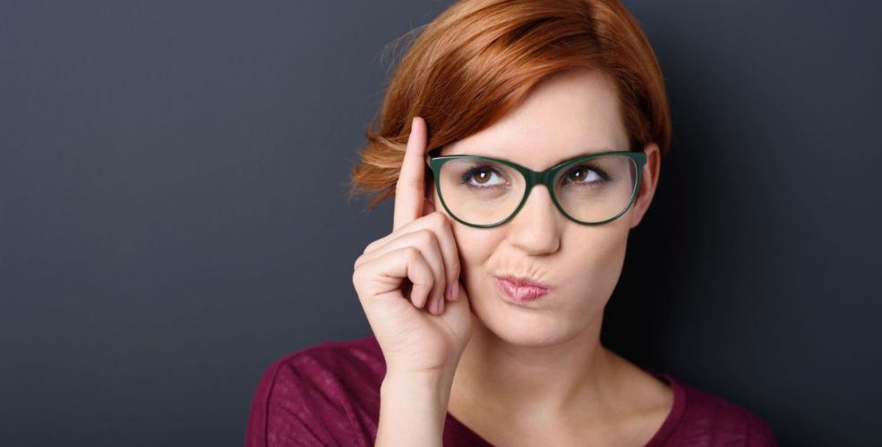 A woman with short red hair and green glasses holding a finger to her temple, looking thoughtful.