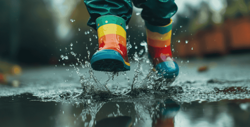 A child wearing rainbow-colored rain boots splashes in a puddle, with water droplets flying around, capturing a moment of carefree joy.