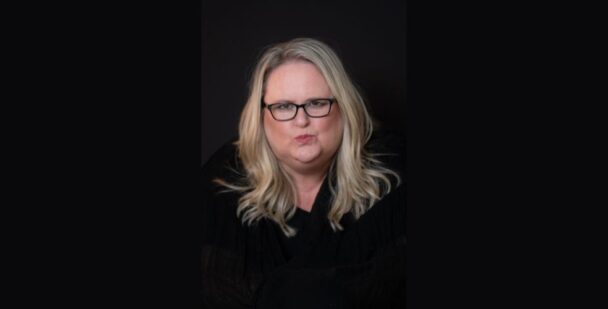 Coach Jasmyne looking directly at the camera, wearing glasses and black, with a confident, thoughtful expression.